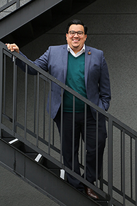 A Latino man standing on steps posing for the camera wearing a gray suit and green sweater