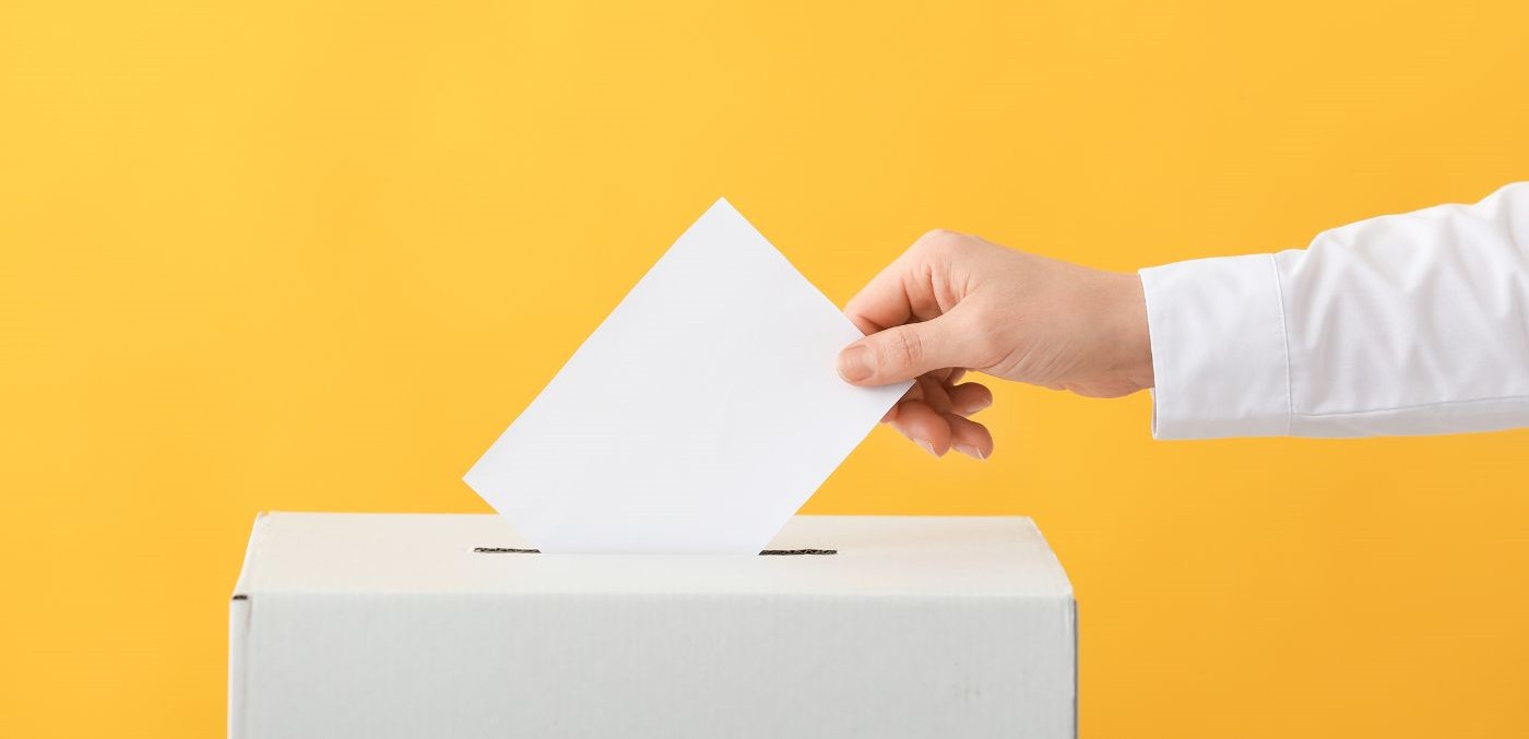 A photo of an arm dropping a ballot into a white box with a yellow background